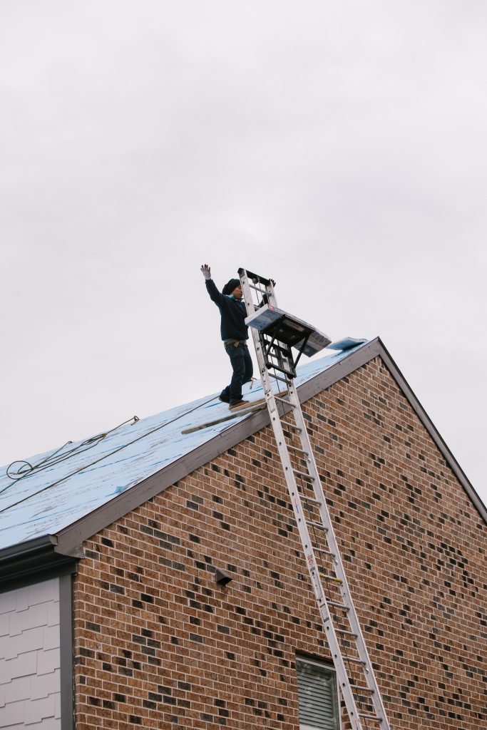 Worker balancing on the edge of a sloped roof with a ladder against a brick house.