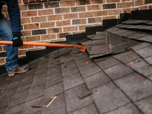 Worker removing shingles from a slanted roof using a long tool near a brick wall.