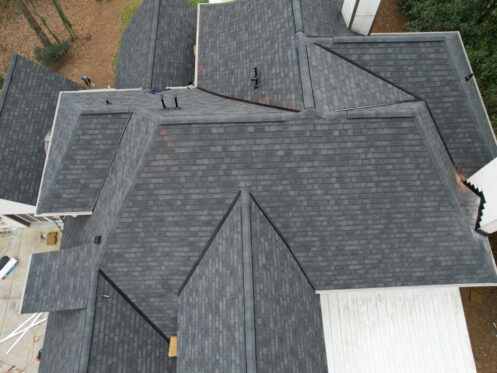 Aerial view of a multi-pitched roof with dark shingles and surrounding trees.