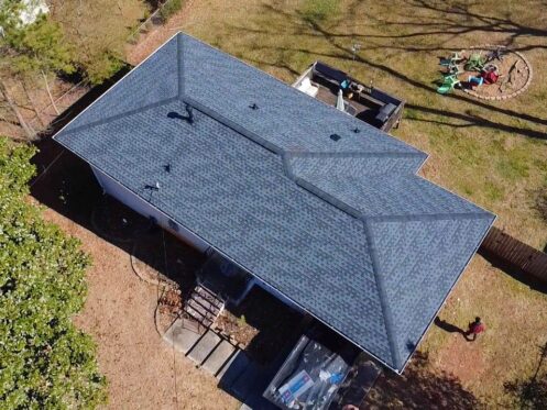 Aerial view of a house with a dark shingle roof and a grassy yard featuring outdoor furniture.
