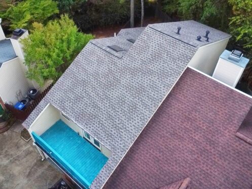 Aerial view of two rooftops, one with gray shingles and the other in reddish tones; green trees nearby.