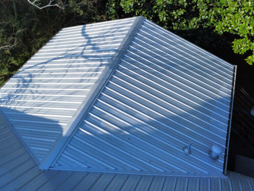 Metal roof viewed from above, featuring ridges and shadows from nearby trees.